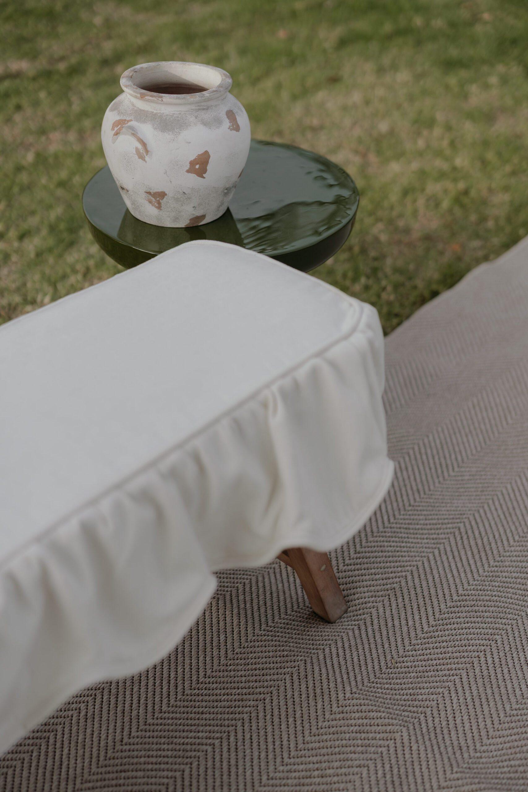Outdoor lounge detail with draped ottoman, green side table, ceramic décor and neutral textured rug by a|CREATE